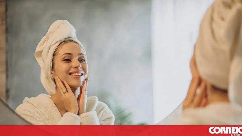 Mulher com toalha na cabeça e roupão, sorri ao espelho