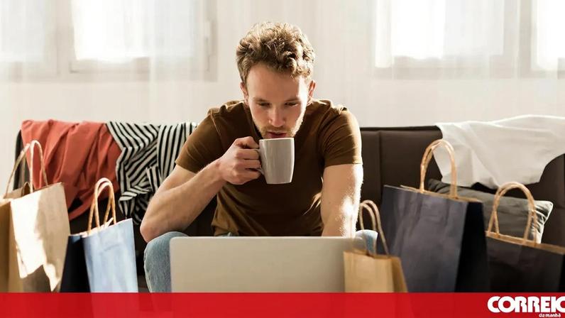 Homem faz compras online com sacos ao lado e bebe uma bebida quente
