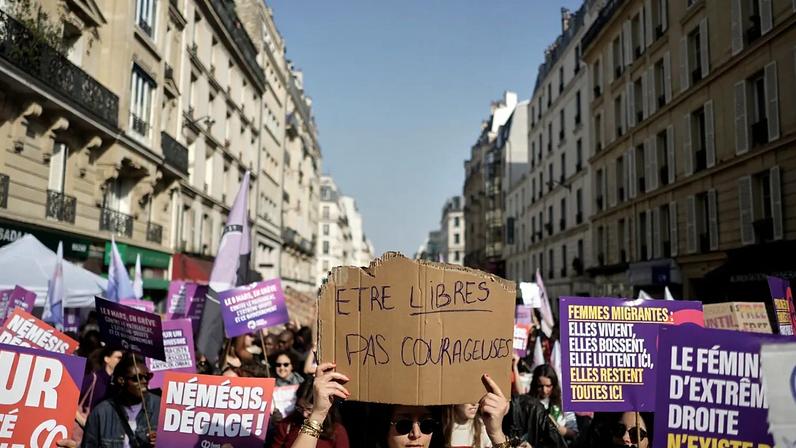 Mulher ao centro exibe cartaz com a frase 'Be free, not brave' durante marcha pelo Dia Internacional da Mulher, em Paris, domingo, 8 de março de 2026