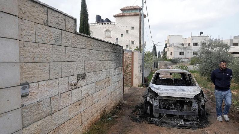 Um homem palestiniano inspecciona um veículo incendiado junto a um graffiti hebraico onde se lê "vingança", na sequência da violência dos colonos israelitas em aldeias da Cisjordânia, 23 de março de 2026.