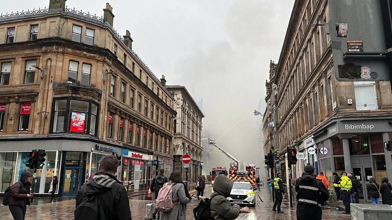 Pessoas observam o fumo que se eleva enquanto bombeiros extinguem os focos restantes de um incêndio que deflagrou num edifício