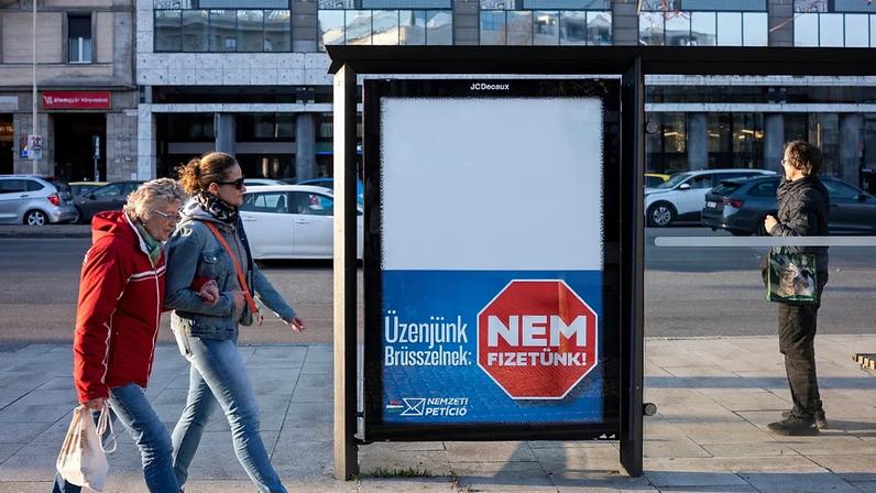 Pessoas caminham junto a um cartaz de campanha que diz: "A nossa mensagem para Bruxelas: Não pagaremos", em Budapeste, Hungria, no sábado, 14 de março de 2026, antes da sessão parlamentar de 12 de abril.