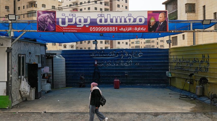 Palestiniana passa por um cartaz com foto de Vladimir Putin e a frase "Lavagem de carros do Putin", em Ramallah, Cisjordânia, 23 de fevereiro de 2026