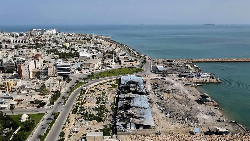 Em pano de fundo os navios no Estreito de Ormuz, danos causados por ataques aéreos vistos num cais do porto da ilha de Qeshm, 13 de abril de 2026