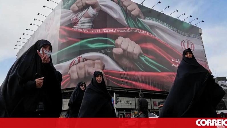 Mulheres iranianas caminham perto de cartaz com a bandeira do Irão, em Teerão