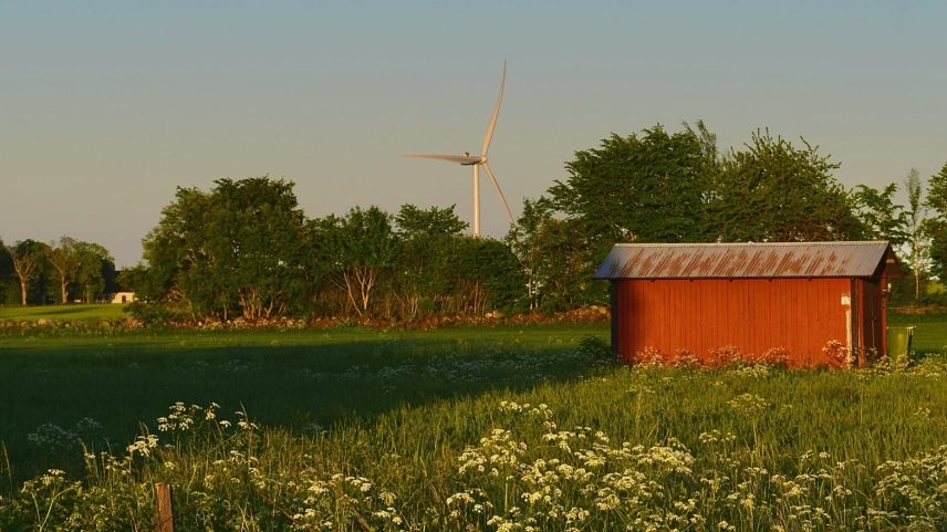 Nesta fotografia, vê-se uma turbina eólica atrás de um barracão vermelho num campo na Suécia