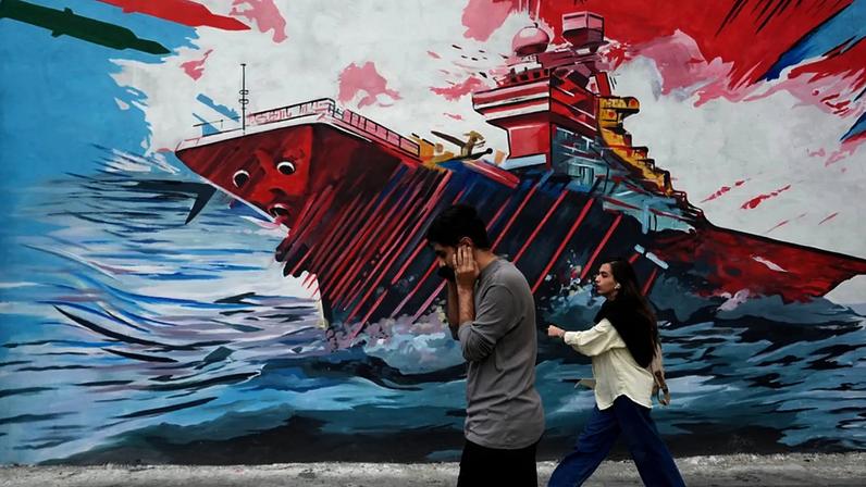 Pessoas passam por um mural que representa um porta-aviões dos EUA sob ataque de mísseis no centro de Teerão, Irão, domingo, 17 de maio de 2026