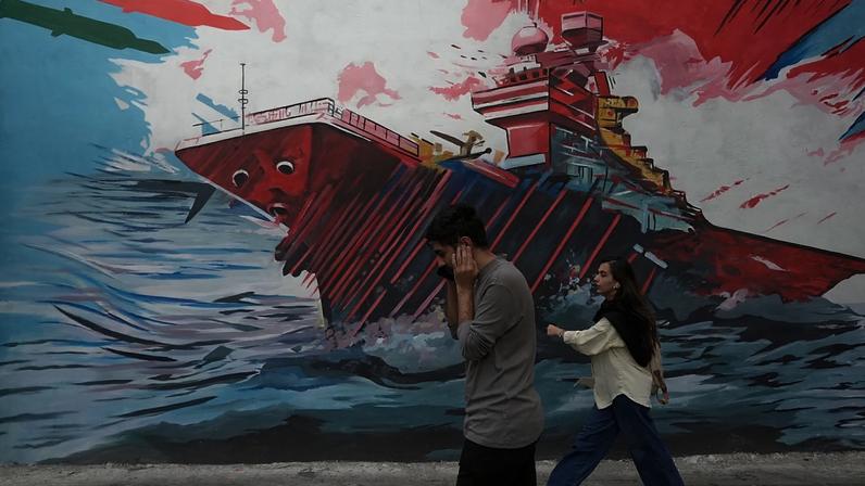 Pessoas passam por mural que mostra um porta-aviões dos EUA sob ataque de mísseis no centro de Teerão, Irão, domingo, 17 de maio de 2026. (AP/Vahid Salemi)