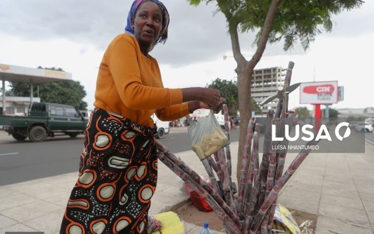 Catarina Floria, uma vendedora de cana-de-açúcar em plásticos nas ruas de Maputo, Moçambique, com cana-de-açúcar ao seu lado, em uma rua movimentada.